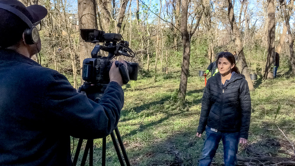 A person being interviewed on camera, standing in the middle of a riparian habitat with trees and vegetation surrounding them.