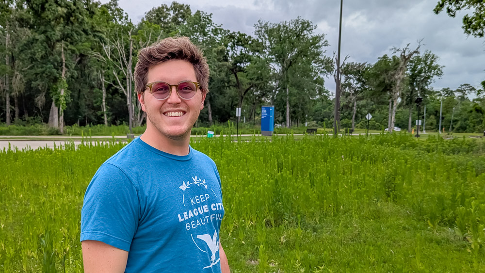 Jeff Fato Smiling person in a blue t-shirt standing in a green field with trees and a cloudy sky in the background.