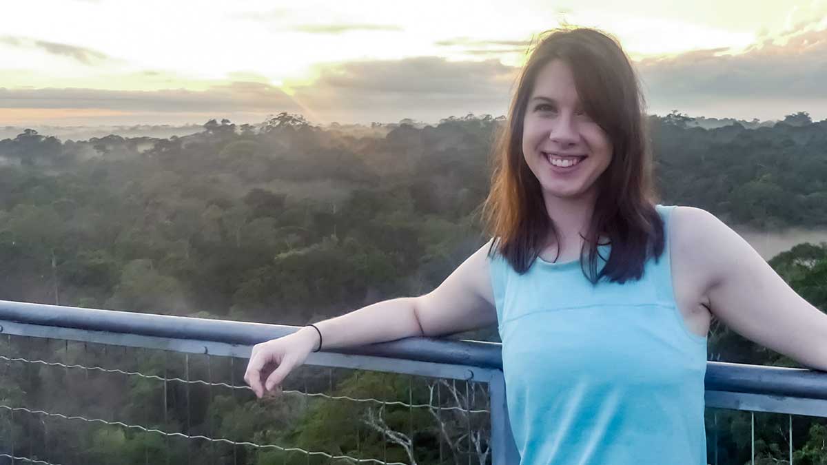 Brandi is smiling at the camera and standing casually against a railing overlooking a forest.