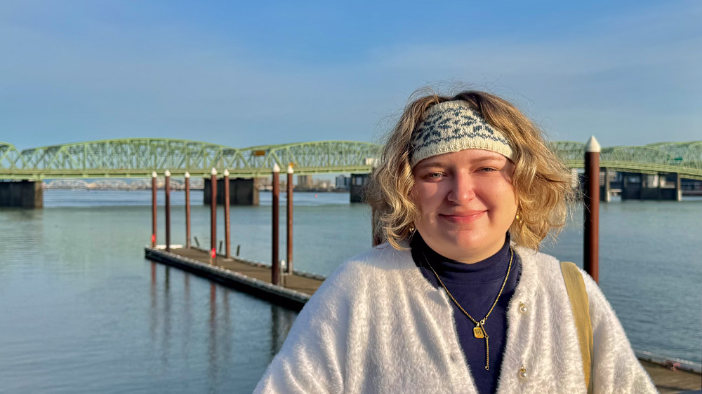 Madeline Sunshine Person smiling near a riverfront dock with a green truss bridge in the background.