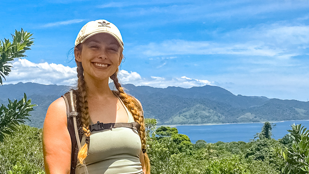 Chelsey is outdoors on a sunny day, mountains and a body of water in the background.